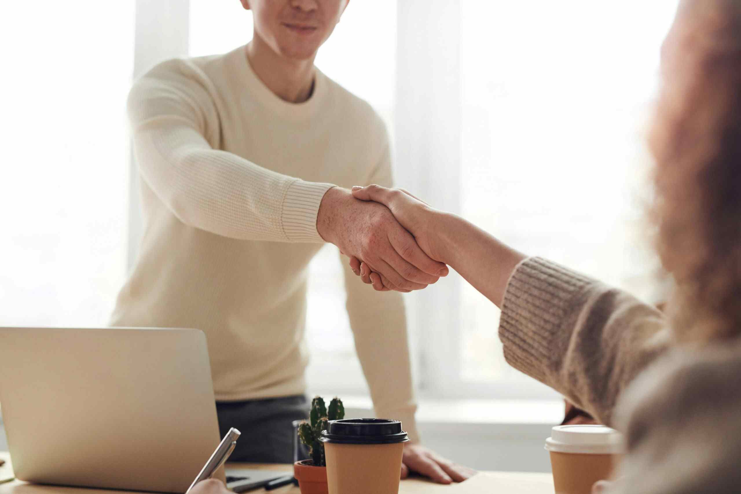 Cierre de trato profesional en una oficina_8_11zon G2 Auto vehículos ocasión - Dos personas estrechándose la mano en una reunión profesional, con una computadora portátil y café en la mesa.