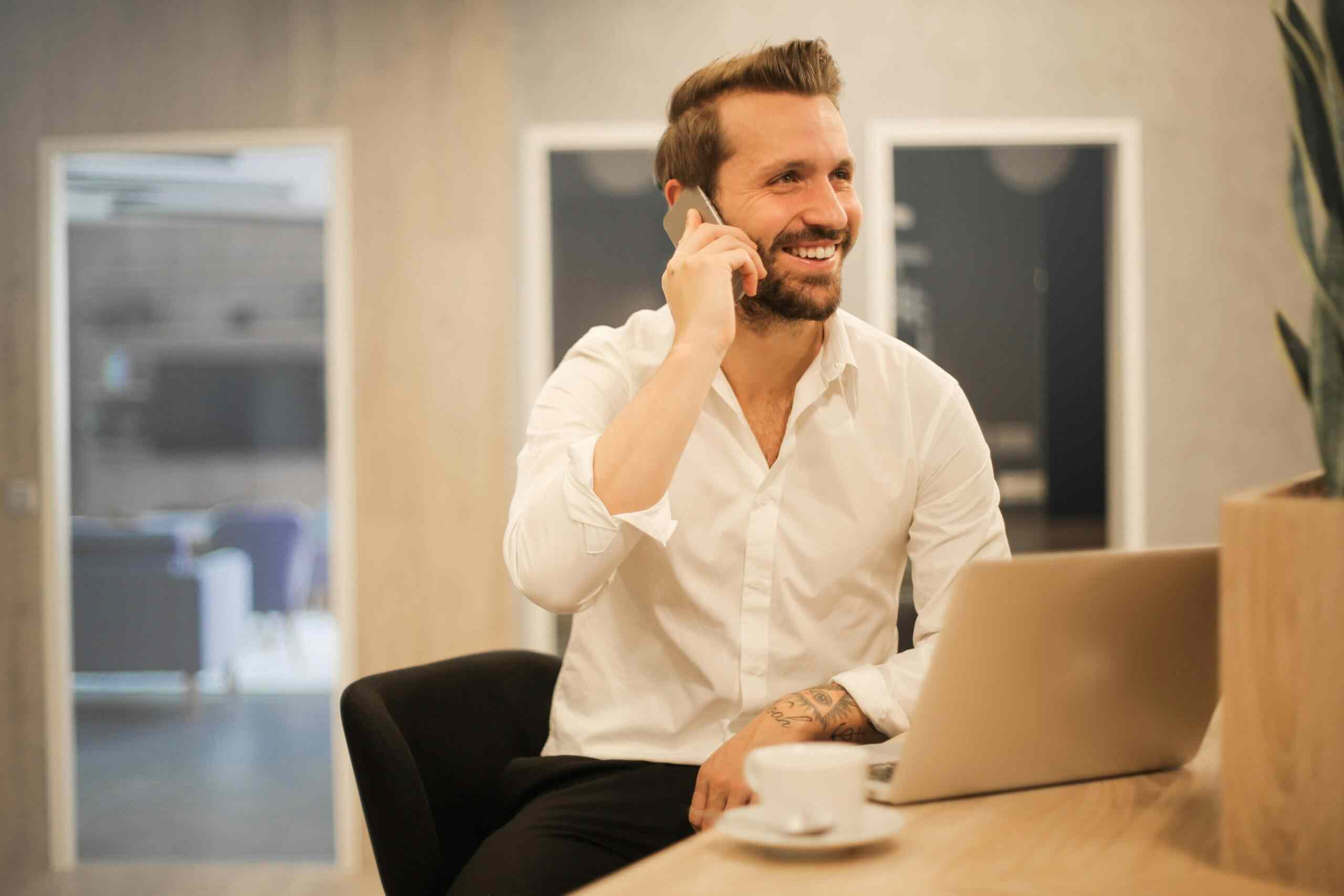 Hombre sonriente en llamada de negocios_9_11zon G2 Auto vehículos ocasión - Hombre sonriente vestido formalmente hablando por teléfono en un espacio de oficina moderno, con una taza de café y un portátil.