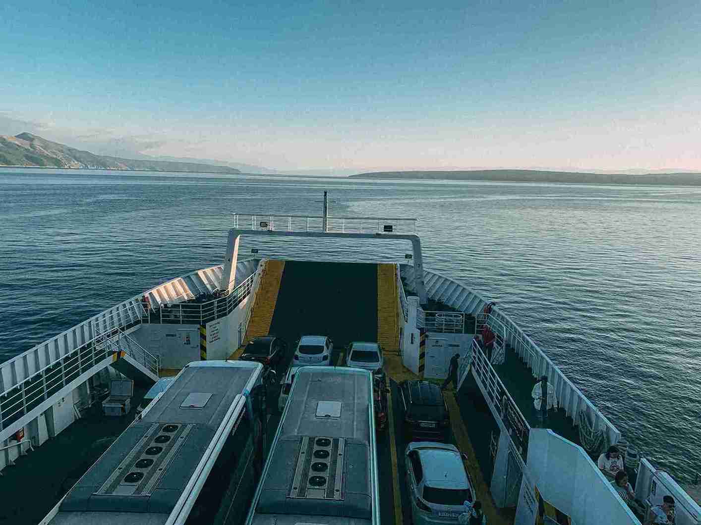 Vehículos Coches en ferry cruzando el océano_12 G2 Auto vehículos ocasión - Camiones y coches a bordo de un ferry navegando en aguas tranquilas, con un paisaje montañoso al fondo.